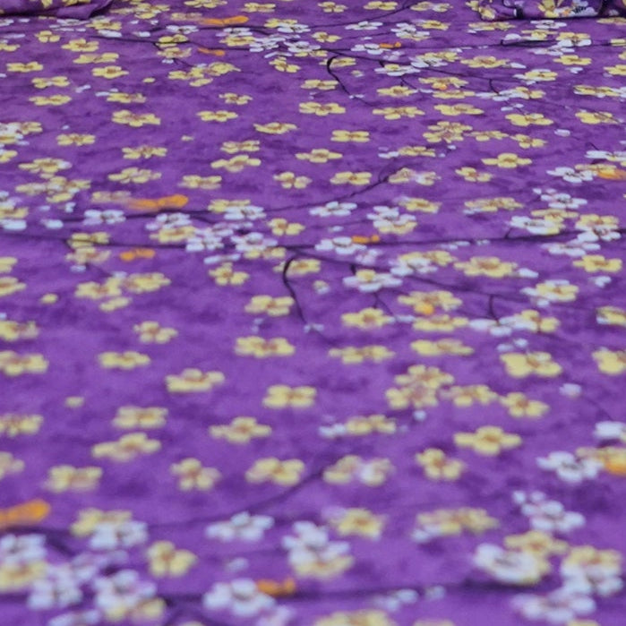 Purple floral comforter set on a bed in a bedroom with white curtains and a lamp.